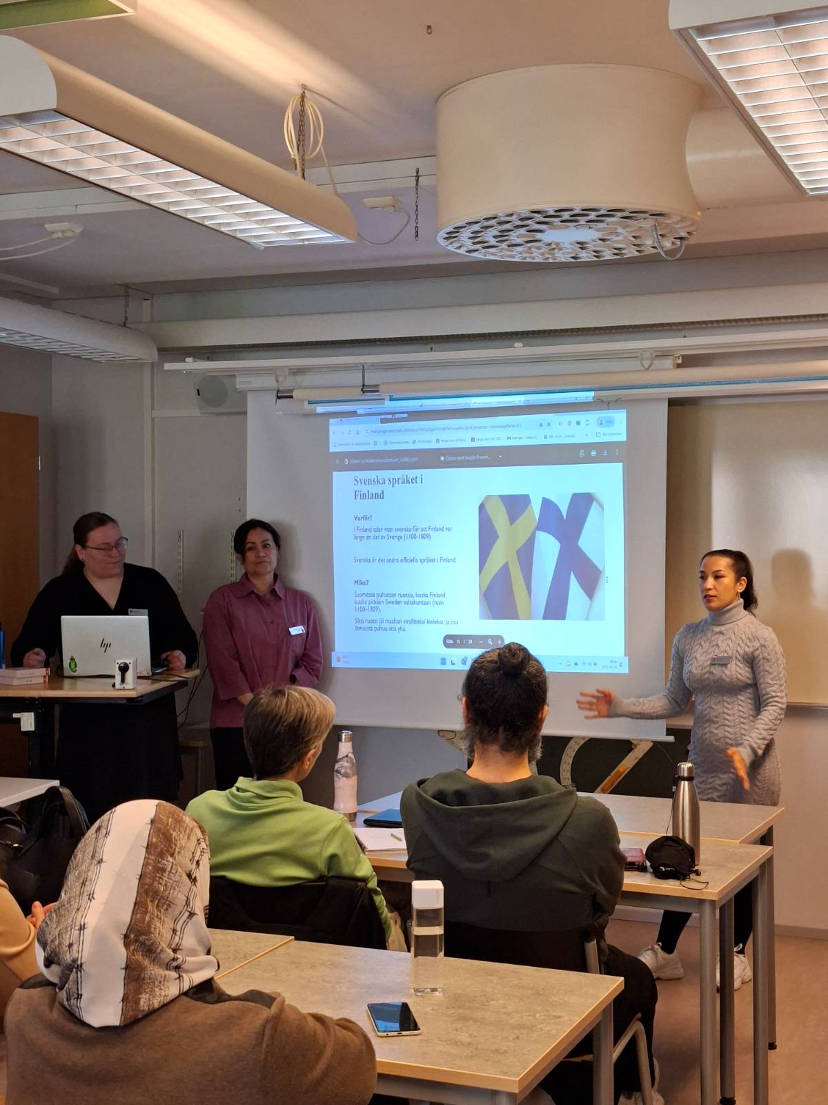Kaksi opiskelijaa esittelee luokan edessä diaa Svenska spåket i Finland. Opettaja seisoo tietokoneen takana sivussa.