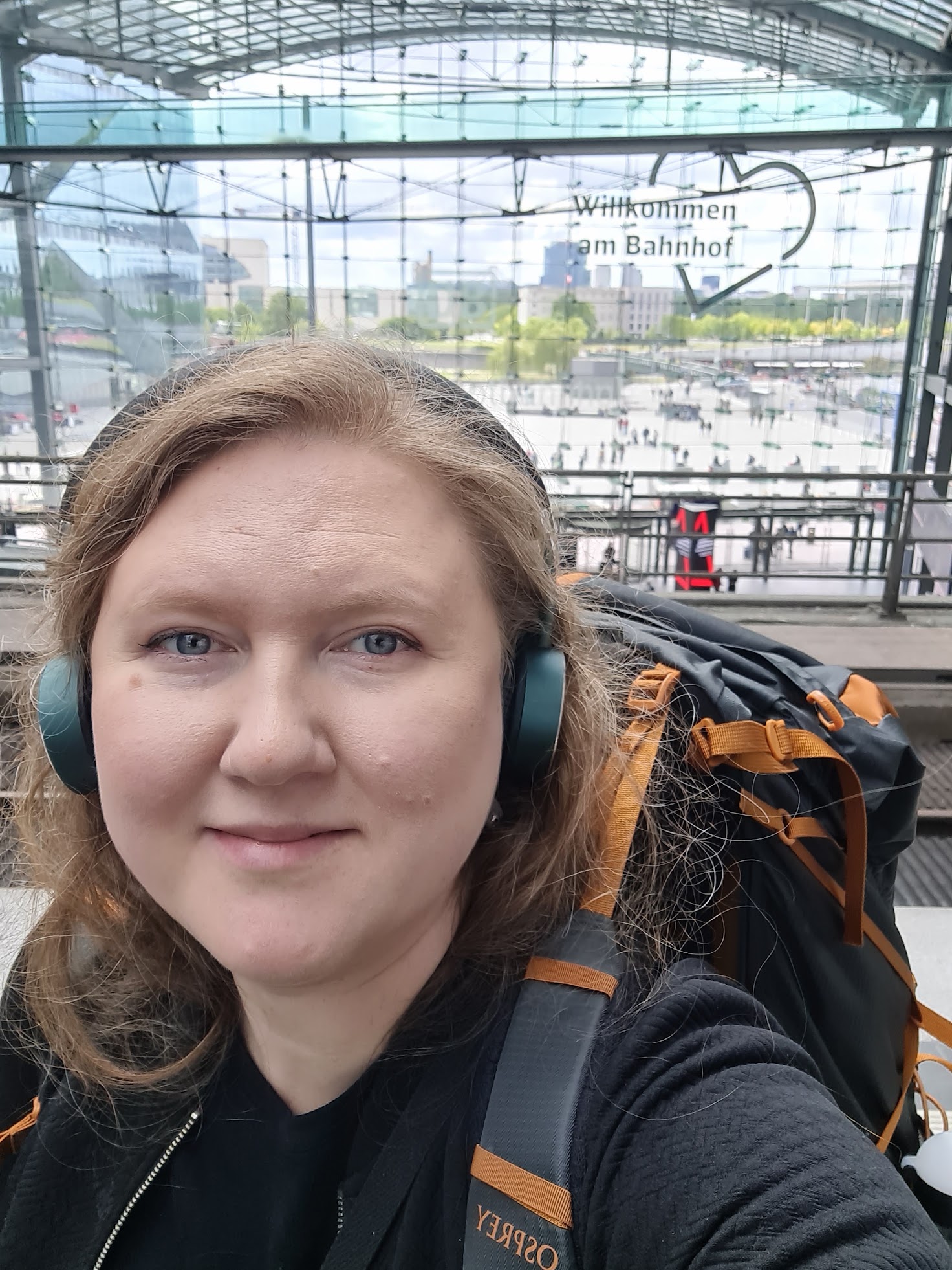 Selfie, jonka taustalla näkyy lasinen asemarakennuksen seinä. Seinällä on sydämen sisällä teksti Willkommen am Bahnhof