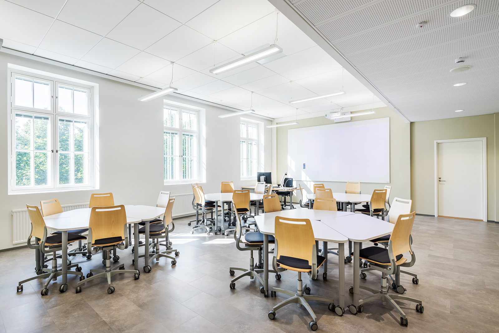 Avara ja valoisa Runeberginkadun ryhmätyötila. Tilassa kolme erillistä pöytäryhmää pyöreässä muodossa. Takana näkyy opettajan pöytä ja whiteboard. Vasemmalta puolelta isot ikkunat tuovat aurinkoa ja valoa luokkaan.