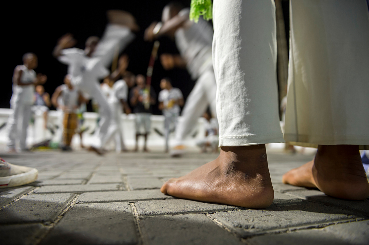 Capoeira Helsinki - kuvassa ryhmä capoeiran harrastajia sumeasti taustalla. Etualalla jalat valkoisissa puuvillahousuissa.